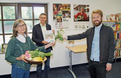 Frau übergibt Rose an Mann, weiterer Mann im Hintergrund vor Bücherregalen