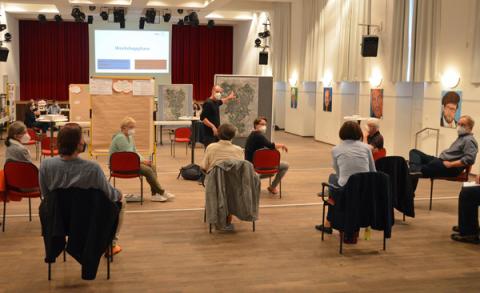 Menschen sitzen auf Abstand, im Hintergrund eine Stellwand mit Luftbild