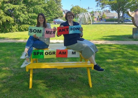 Zwei Frauen sitzen auf Picknickbank und halten Schriftzug hoch