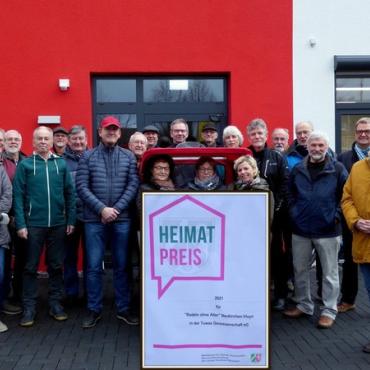 Menschen sitzen in Fahrradrikscha, andere stehen darum herum, im Vordergrund Plakat mit Heimatpreis-Auszeichnung Menschen sitzen in Fahrradrikscha, andere stehen darum herum, im Vordergrund Plakat mit Heimatpreis-Auszeichnung